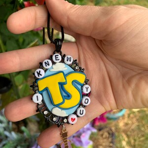 May include: An oval pendant necklace featuring a black filigree frame with small clear rhinestones. The center has a light blue background with white clouds and large yellow letters reading TS. White letter beads spell I KNEW YOU around the edge.