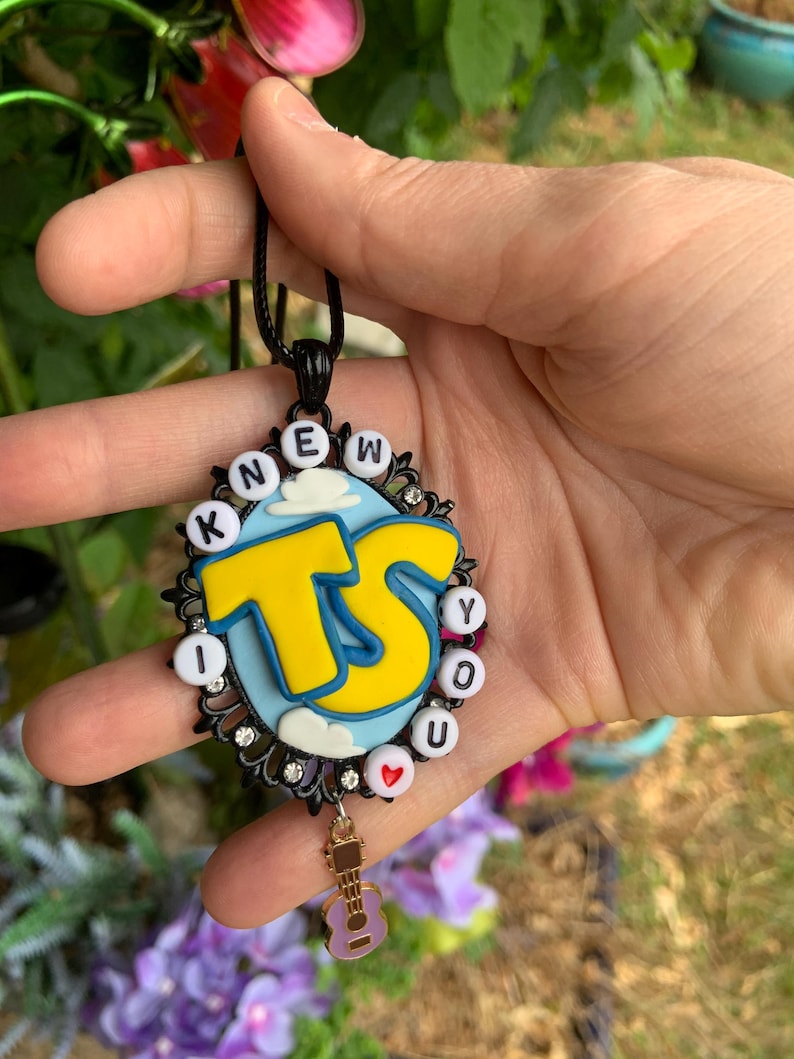May include: An oval pendant necklace featuring a black filigree frame with small clear rhinestones. The center has a light blue background with white clouds and large yellow letters reading TS. White letter beads spell I KNEW YOU around the edge.