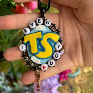 May include: An oval pendant featuring a light blue background with white clouds and large yellow letters TS. The border is black with small clear rhinestones and white beads spelling KNEW YOU with a red heart. A small purple guitar charm hangs below.