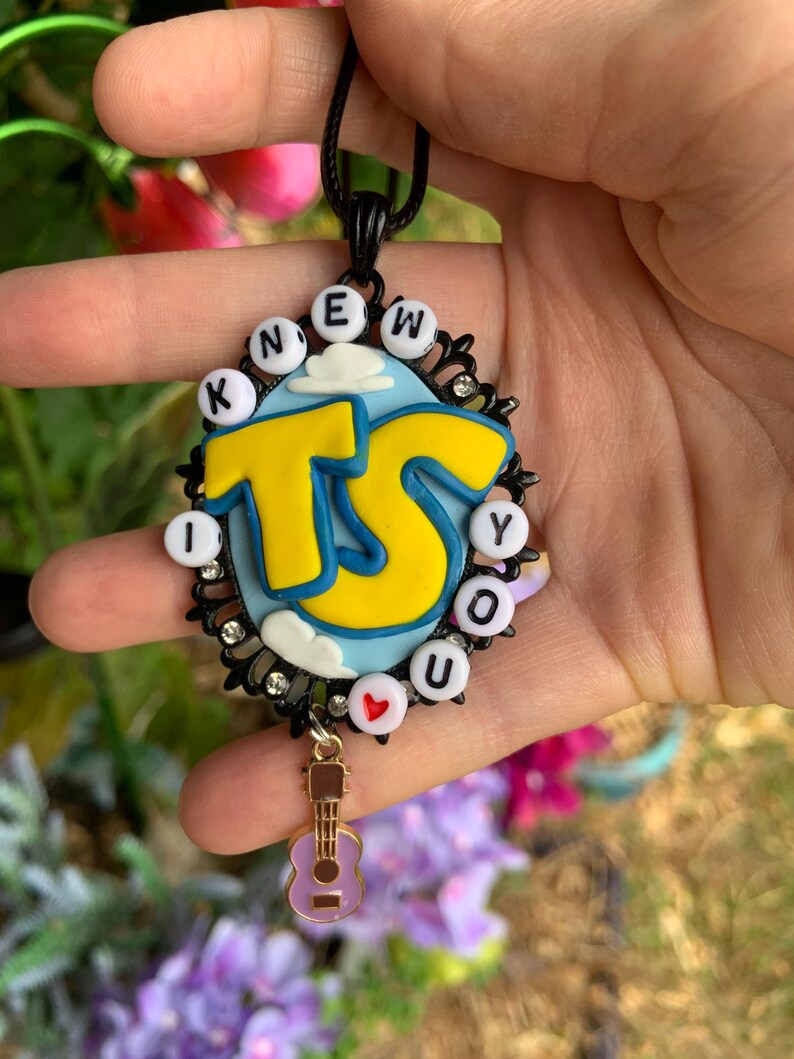 May include: An oval pendant featuring a light blue background with white clouds and large yellow letters TS. The border is black with small clear rhinestones and white beads spelling KNEW YOU with a red heart. A small purple guitar charm hangs below.