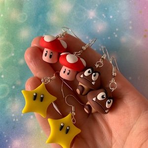 Puede incluir: Pendientes hechos a mano con personajes de un videojuego popular. Los pendientes presentan diseños de champiñones rojos y blancos, Goombas marrones y estrellas amarillas, todos con detalles en blanco y negro y ganchos plateados.