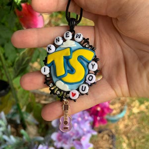 May include: A black oval pendant featuring a light blue sky background with white clouds and large yellow letters reading TS. The border is adorned with white letter beads spelling I KNEW YOU and a small red heart bead, plus a dangling light purple guitar charm.