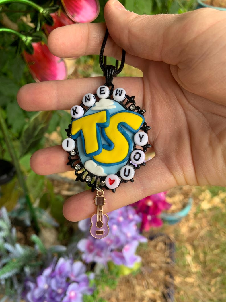May include: A black oval pendant featuring a light blue sky background with white clouds and large yellow letters reading TS. The border is adorned with white letter beads spelling I KNEW YOU and a small red heart bead, plus a dangling light purple guitar charm.