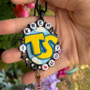 May include: An oval pendant on a black cord featuring a light blue sky background with white clouds and large yellow letters TS. The border is black with small clear rhinestones and white beads spelling KNEW YOU with a red heart. A small guitar charm hangs below.