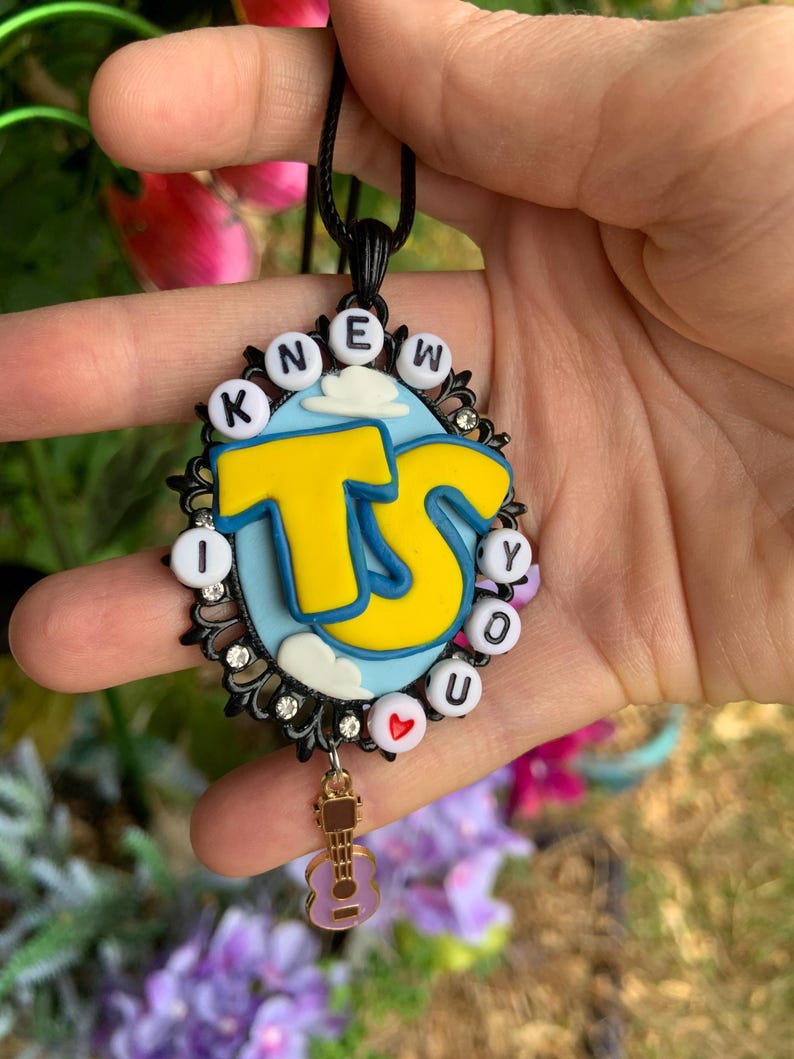 May include: An oval pendant on a black cord featuring a light blue sky background with white clouds and large yellow letters TS. The border is black with small clear rhinestones and white beads spelling KNEW YOU with a red heart. A small guitar charm hangs below.