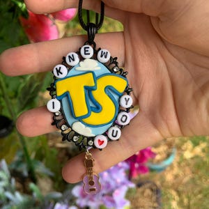 May include: A black oval pendant featuring a light blue background with white clouds and large yellow letters TS. The border is decorated with white letter beads spelling I KNEW YOU and a small red heart bead, plus a dangling gold guitar charm.
