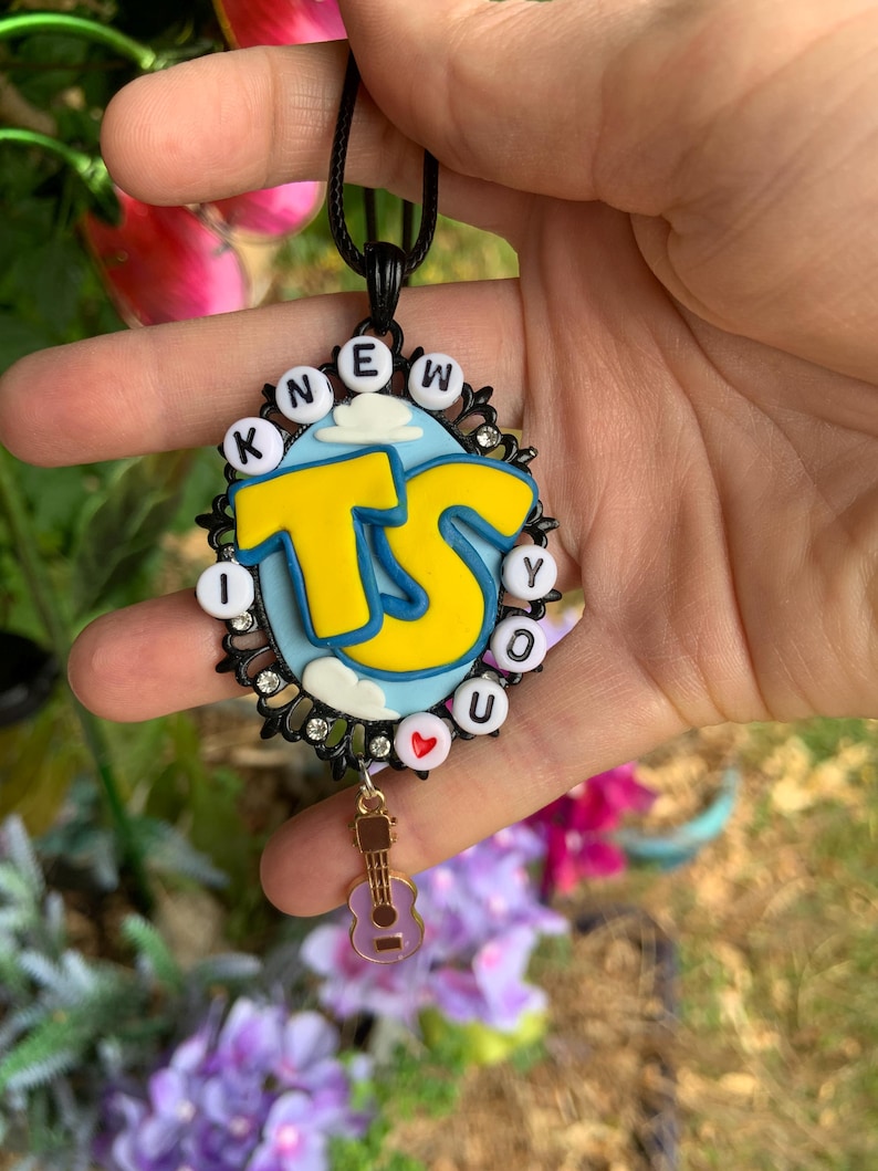 May include: A black oval pendant featuring a light blue background with white clouds and large yellow letters TS. The border is decorated with white letter beads spelling I KNEW YOU and a small red heart bead, plus a dangling gold guitar charm.