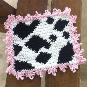 May include: A crocheted square with a black and white cow print design. The square has a pink crocheted border.