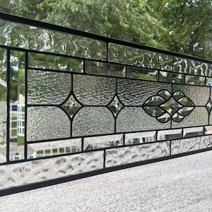May include: A rectangular stained glass window panel featuring a black metal frame. The design incorporates clear textured glass segments, beveled glass accents, and a central decorative motif composed of interlocking geometric shapes.