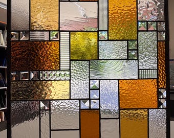 May include: A rectangular stained glass panel featuring a geometric grid of various glass textures and colors. The composition includes clear, amber, and yellow glass panes with rippled, pebbled, and smooth finishes, all joined by dark metal cames.