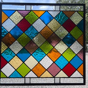May include: A square stained glass panel featuring a diamond grid pattern with various colored glass panes including blue, green, orange, red, pink, and clear textured glass, all encased within a black metal frame with vertical accents.
