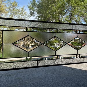 May include: Rectangular stained glass window with a black metal frame. The window features clear textured and diamond-shaped glass panels. The glass reflects the outdoor surroundings, creating a unique visual effect. The window is suspended by a chain.