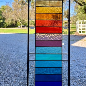 May include: A rectangular stained glass panel featuring a vertical arrangement of horizontal glass strips in a rainbow spectrum of colors including yellow, orange, red, purple, teal, and blue, framed by a black metal border.