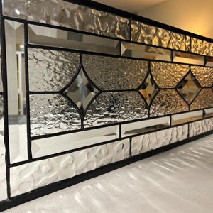 May include: A rectangular stained glass panel with a black frame. The panel features clear, textured, and mirrored glass pieces arranged in geometric patterns, including diamonds and rectangles. The glass reflects light, creating a shimmering effect.