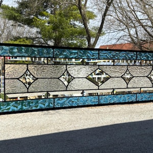 May include: Rectangular stained glass window panel with a black frame. The panel features a central design of clear and textured glass with diamond-shaped accents, bordered by turquoise textured glass. The panel is suspended by a chain.