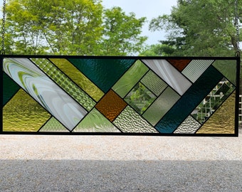 May include: A stained glass window with a geometric pattern in shades of green, gold, and brown. The window is framed in black and hangs from a chain.