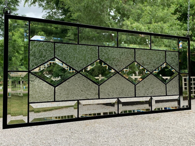 Pu&ograve; includere: Una finestra in vetro colorato con un design geometrico che presenta una fila di forme a diamante al centro. Il vetro &egrave; trasparente e testurizzato, con linee di piombo nere che delineano il design.