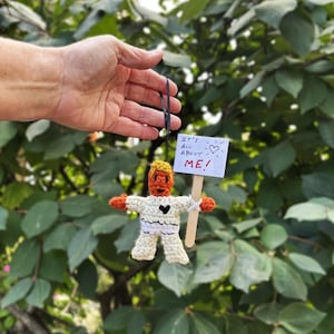 May include: A handmade crochet doll with orange hair and arms, wearing a white outfit with a black heart. The doll holds a sign that reads "IT'S ALL ABOUT ME!" with a heart symbol. The doll is suspended by a black string.