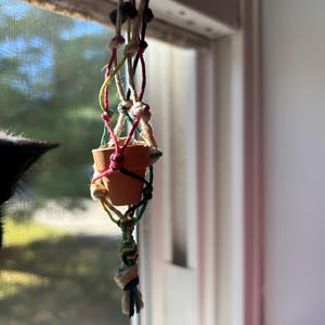 May include: A miniature terracotta plant pot hanging from a macramé plant hanger. The hanger is made of natural and coloured cords, including red, green, yellow, and black. The pot is empty and hangs near a window.