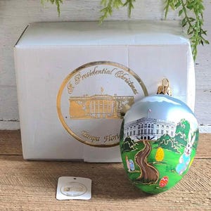 Puede incluir: Un adorno navideño blanco y plateado con una escena pintada en verde, blanco y azul de la Casa Blanca y conejos de Pascua. El adorno está en su caja original, que tiene un sello dorado con el texto "The Presidential Christmas, Tampa Florida".
