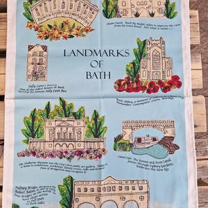 May include: A blue tea towel with illustrations of Bath landmarks, including the Royal Crescent and Pultney Bridge. The towel has text describing each landmark, such as Sham Castle and Bath Abbey. The words "Landmarks of Bath" are in the center of the design.