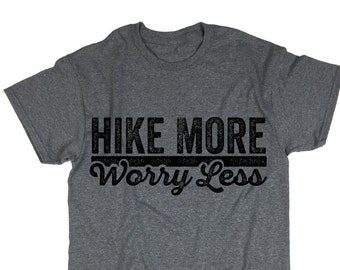 May include: A grey t-shirt featuring the black text "HIKE MORE Worry Less". The text is centered on the front. The shirt has short sleeves and a crew neck. The fabric appears to be a heathered blend.