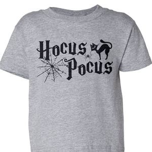 K&ouml;nnte beinhalten: Ein graues T-Shirt mit kurzen &Auml;rmeln mit dem Text "Hocus Pocus" in Schwarz, mit einer schwarzen Katze und einem Spinnennetz.