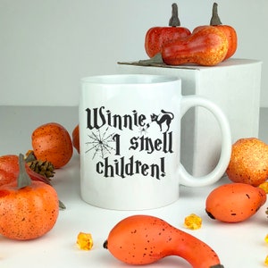 May include: White ceramic mug with a black and white graphic that says "Winnie, I smell children!" with a spider and a black cat. The mug is surrounded by orange pumpkins.