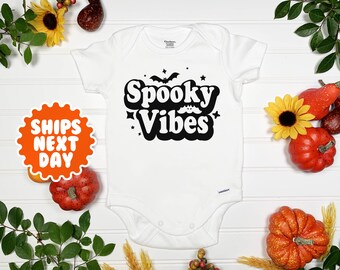 May include: White baby onesie with black text that reads "Spooky Vibes" in a retro font. The text is surrounded by stars and bats.