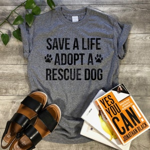 May include: Gray t-shirt with the text "SAVE A LIFE *ADOPT A* RESCUE DOG" in black lettering. The shirt is on a wooden surface with a pair of brown sandals and a stack of books.