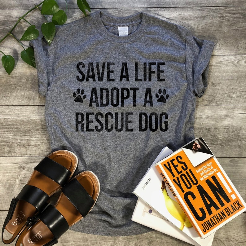 May include: Gray t-shirt with the text "SAVE A LIFE *ADOPT A* RESCUE DOG" in black lettering. The shirt is on a wooden surface with a pair of brown sandals and a stack of books.