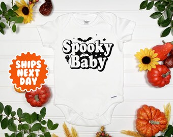 May include: White baby onesie with black text that reads "Spooky Baby" in a retro font. The text is surrounded by stars and bats.