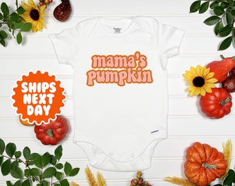 May include: White baby onesie with the text "mama's pumpkin" in pink and orange letters. The onesie is surrounded by autumnal decorations, including pumpkins and sunflowers.