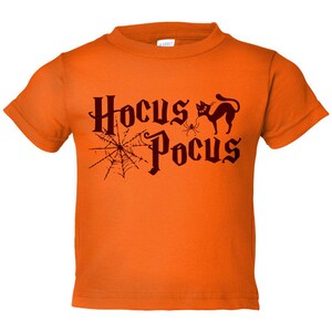 K&ouml;nnte beinhalten: Orangefarbenes T-Shirt mit kurzen &Auml;rmeln und dem Text "Hocus Pocus" in schwarzer Schrift. Auf dem Shirt sind au&szlig;erdem eine schwarze Katze und eine Spinne abgebildet.