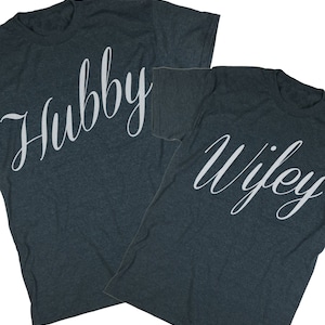 Puede incluir: Dos camisetas de color gris oscuro jaspeado con texto blanco. Una camiseta dice "Hubby" y la otra dice "Wifey".