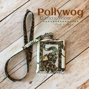 Può includere: Un porta badge in tessuto marrone e verde con una finestra in plastica trasparente. Il porta badge presenta un design a forma di elefante e il testo "Pollywog ID badge holder Needle & Anchor SUPPLY CO".