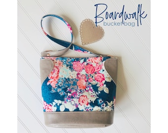 May include: A floral patterned bucket bag with a brown leather strap and trim. The bag has a brown leather heart-shaped tag attached to the strap. The text "Boardwalk bucket bag" is written in white on a black background.