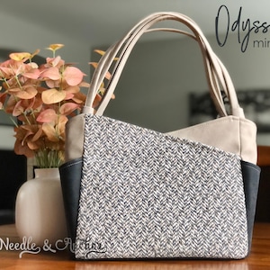Könnte beinhalten: Eine beige und schwarze Mini-Tragetasche mit Fischgrätenmuster. Die Tasche hat einen oberen Griff und einen Reißverschluss. Die Tasche ist mit "Odyssey mini tote" und "Needle & Anchor" beschriftet.