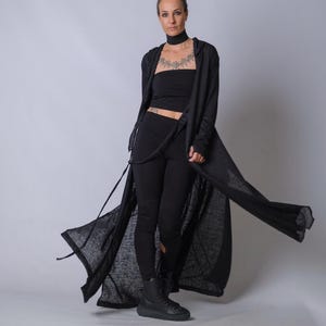 Könnte beinhalten: Eine Frau in einem schwarzen Outfit, bestehend aus einem langen, fließenden Cardigan, einem Bandeau-Top, Leggings und Stiefeln. Der Cardigan hat eine Kapuze und Bindebänder. Das Outfit ist ganz schwarz und erzeugt einen schlanken, monochromen Look. Die Frau steht vor einem schlichten Hintergrund.