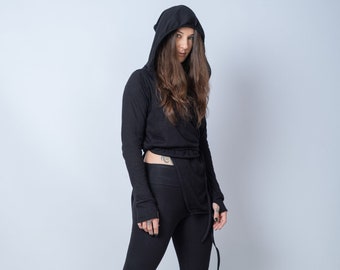 May include: A woman wearing a black hooded top and black leggings. The top has a long, flowing design and the leggings are form-fitting. She is wearing black sneakers with white soles.