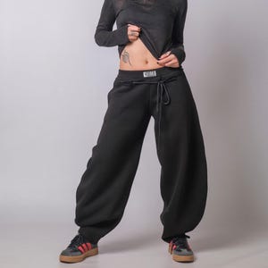 Op de afbeelding: Een persoon draagt een donkergrijs top met lange mouwen en een zwarte joggingbroek. De joggingbroek heeft een trekkoord en een witte band met het woord "YOGTS" erop gedrukt. De persoon draagt ook sneakers.