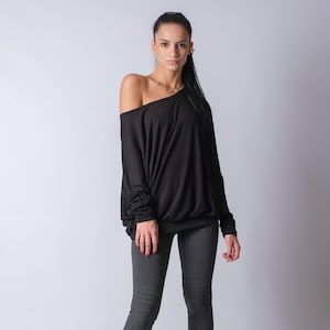 Könnte beinhalten: Eine Frau trägt ein schwarzes, langärmeliges Shirt mit offener Schulter, graue Leggings und schwarze Stiefeletten.