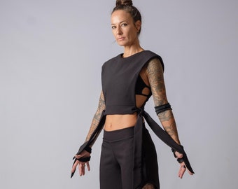 May include: A black two-piece set consisting of a sleeveless cropped top with wide shoulders and side tie closures, paired with matching mid-calf length trousers featuring a circular cutout on the left leg and black slide sandals.