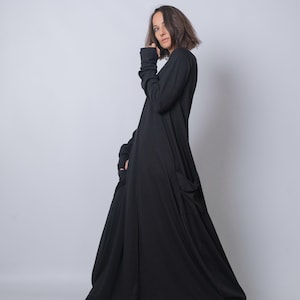 Puede incluir: Vestido negro largo y fluido con mangas largas y bolsillos grandes. El vestido tiene un corte holgado y un diseño sencillo. La modelo lleva zapatos negros. El fondo es blanco liso.