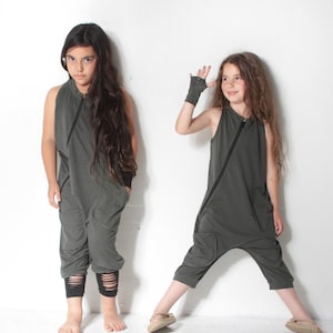 May include: Two young people wearing matching olive green sleeveless jumpsuits with black accents. The jumpsuits have a zipper down the front and a loose, comfortable fit. The person on the left is standing with their arms at their sides, while the person on the right is standing with one arm raised and their hand in a fingerless glove.