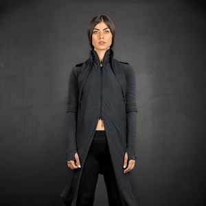 May include: A long, dark grey hooded jacket with a zip closure. The jacket has a high collar and a long, flowing hem. The model is wearing black leggings and black trainers.