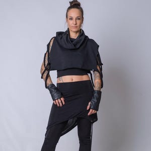 May include: Black outfit featuring a hooded top with cut-out sleeves, a crop top, a draped skirt, and leggings. The outfit includes black leather gloves and boots. The model is standing in front of a gray background.