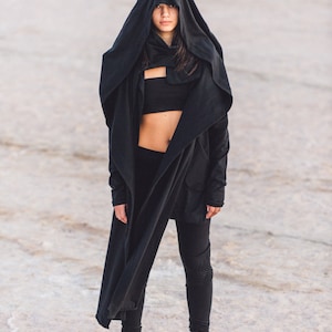 May include: A person wearing a black hooded jacket, black leggings, and black and white sneakers. The person is standing on a white, sandy surface.