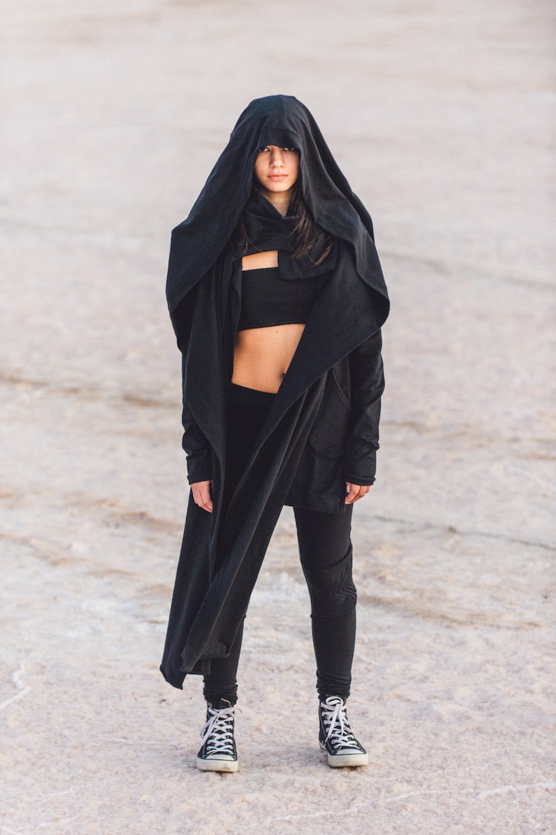 May include: A person wearing a black hooded jacket, black leggings, and black and white sneakers. The person is standing on a white, sandy surface.