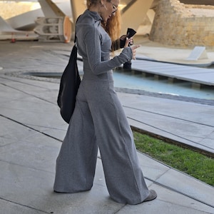 May include: A woman wearing a gray ribbed turtleneck top and matching wide-leg pants. She is standing on a concrete surface with a black bag slung over her shoulder.